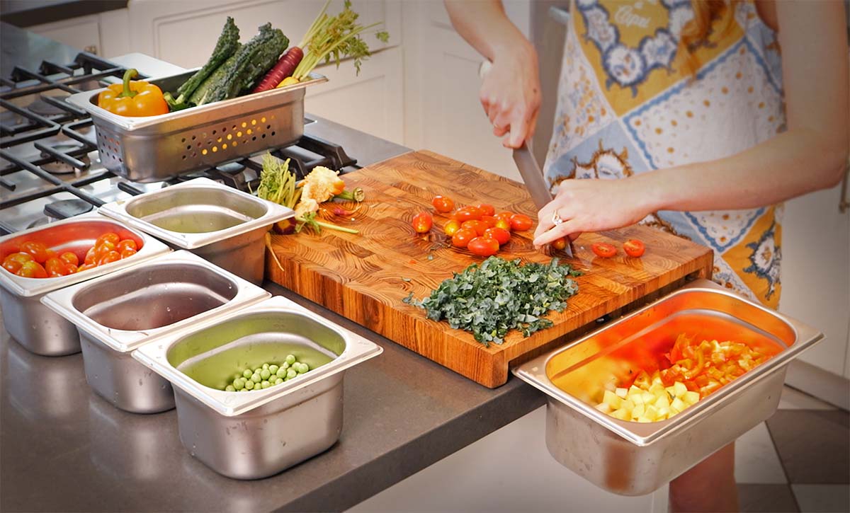 Image of the Good Board in action, with a person adeptly chopping and preparing different vegetables, using several stainless steel containers together with the teak cutting board.